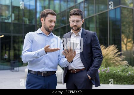 Two professional businessmen collaborating and discussing a new project on a digital tablet, standing confidently outside a modern corporate building, representing teamwork and innovative solutions Stock Photo