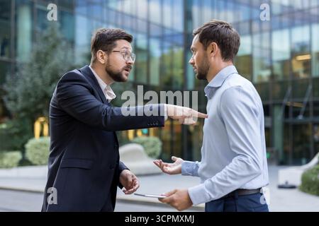 Two angry businessmen are having a tense argument, one pointing a finger while the other gestures in disagreement, depicting workplace stress and conflict outside a modern office building Stock Photo