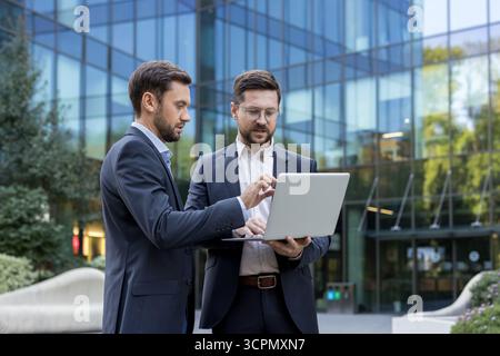 Two businessmen stand outside a glass office building, collaborating over a laptop as they review data and plan strategy, confident and focused in an urban corporate setting Stock Photo
