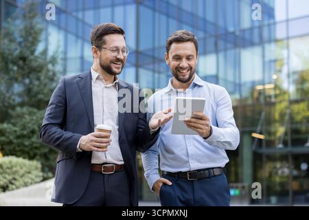 Two cheerful male colleagues brainstorming and collaborating on a new business project using a digital tablet while standing outdoors in front of a modern office building Stock Photo