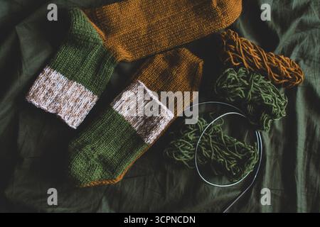 Cozy knitted socks with yarn ball and knitting needles. Striped wool socks on dark green sheet. Cozy autumn colours. Woven crochet close up. Stock Photo