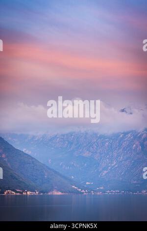 Calm pre-dawn over Kotor Bay, Montenegro, with misty mountains, soft clouds, and pink-blue sky reflecting in still waters before sunrise. Stock Photo