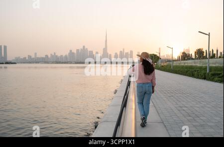 Dubai skyline silhouette at sunset with a woman wearing a sunhat and shirt standing at the waterfront promenade Stock Photo