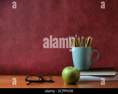 Vintage study desk setup with copy space - Glasses, apple, and pencils in a textured mug on a book stack and wooden desk. Educational concept with red Stock Photo