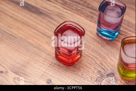 Three square-shaped glasses filled with colorful beverages and ice cubes, placed on a rustic wooden surface. The primary focus is on the red drink. Stock Photo