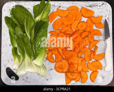 Sliced carrots in half moons, and cleaned fresh bok choy rest on a white cutting board with a paring knife. Stock Photo