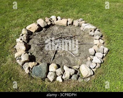 A round backyard fire pit circled by small rocks in a grassy lawn with small sticks as kindling is ready for lighting. Stock Photo