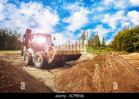 Power shovel stands in working position close to large sand heap, bucket touching granular surface while fresh soil falls from edge. Stock Photo