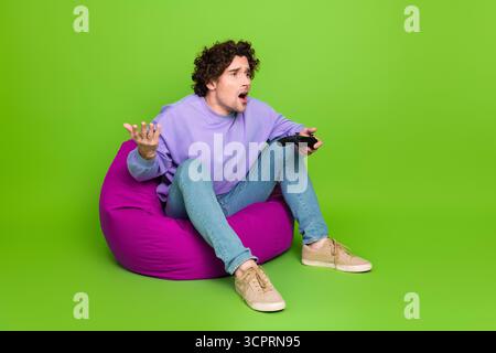 Funny man with curly hair seated on a colorful bean bag holding a gaming controller on a vibrant green background Stock Photo