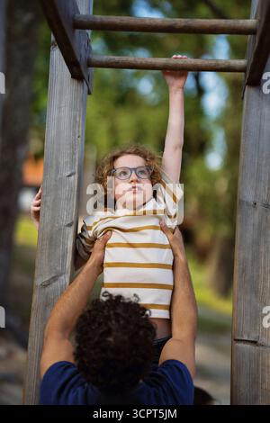 Male guide helping boy in hanging on monkey bars at summer camp Stock Photo