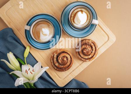 Two cups of cappuccino with foam art and cinnamon rolls on wooden board with lily flower. Flat lay breakfast composition, top view. Stock Photo