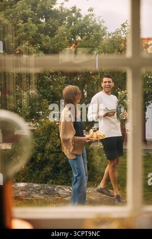 Carefree man enjoying with female friend holding plate of bread while walking in back yard seen from window Stock Photo