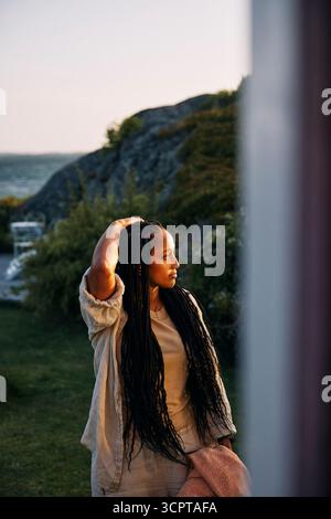 Contemplative woman with hand in hair standing in back yard of vacation home seen from window Stock Photo