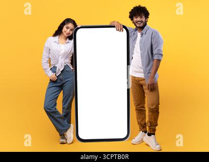 Excited young indian man and woman leaning on giant smartphone with mockup, promoting app or website Stock Photo