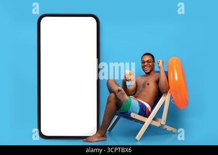Excited black guy with tropical cocktail chilling in lounge chair near huge smartphone with blank screen for your ad Stock Photo