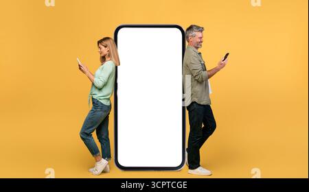 Middle aged man and woman using smartphones, leaning on large phone with empty screen, advertising website, mockup Stock Photo