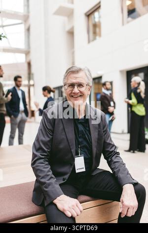 Portrait of smiling confident senior businessman sitting on bench in convention center Stock Photo