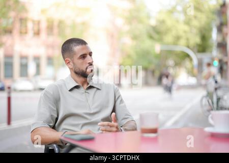 A man sits at a table with a cup of coffee in front of him. He is wearing a shirt and has a beard. The scene is set in a city with a few cars and bicy Stock Photo