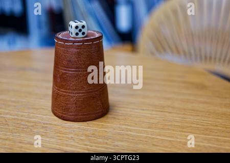 A leather dice cup with white stitching stands upside down on a wooden table. A single die rests on its base showing five on top with four and six on Stock Photo