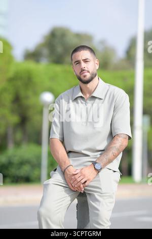 A man in a grey shirt and khaki pants sits on a curb. He is wearing a watch and has tattoos on his arms Stock Photo