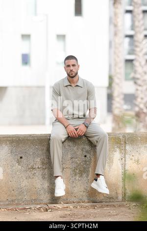 A man is sitting on a wall with his legs crossed. He is wearing a white shirt and khaki pants Stock Photo