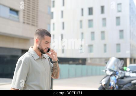 A man is talking on his cell phone while walking down the street. He is wearing a grey shirt and has tattoos on his arm. The scene is set in a city wi Stock Photo