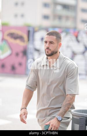 A man in a tan shirt is walking down a street with graffiti on the wall behind him. He is wearing a watch and has tattoos on his arms Stock Photo