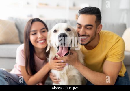 Portrait of happy multiracial couple hugging adorable dog, cuddling with their pet at home Stock Photo
