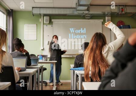 Rear view of teenage female student with hand raised asking question to professor during Q and A session in classroom Stock Photo
