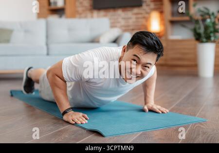 Strong asian mature man making strength workout, standing in plank pose or doing push ups at home on yoga mat Stock Photo
