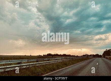 Rural Road under Stormy Sky with Sunlight Breaking Through Clouds in Scenic Landscape Stock Photo