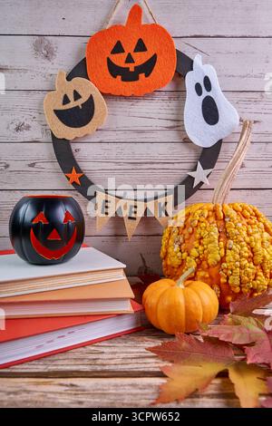 Halloween still life with felt wreath (jack‑o’‑lanterns and ghost), books, warted and mini pumpkins, walnuts, and autumn leaves on rustic wood. Stock Photo