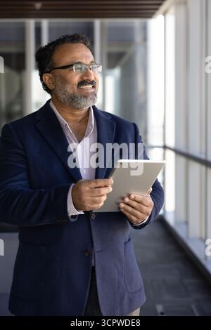 Businessman holds digital tablet, looks out window with thoughtful expression Stock Photo