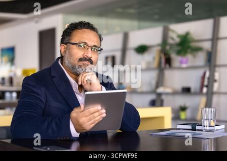 Serious Indian businessman holding tablet seated at modern office table Stock Photo