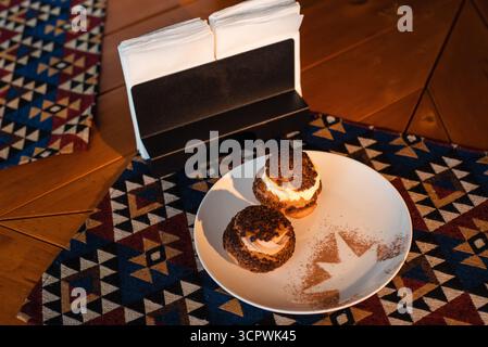 Two cream puffs with chocolate topping on a white plate with cocoa powder design, placed on a wooden table with geometric-patterned placemats and a na Stock Photo