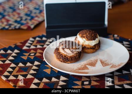Two cream puffs on a white plate with a geometric-patterned tablecloth. Stock Photo