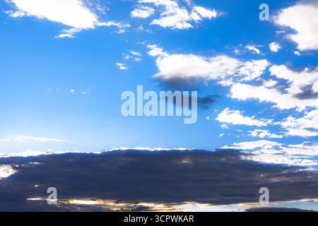 Blue sky is adorned with fluffy white and dark grey clouds, indicating varying weather or light conditions. Below, a band of darker clouds Stock Photo