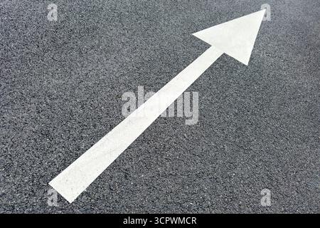 White directional arrow painted on dark asphalt, indicating forward movement or guidance, often seen on roads, parking lots, or pathways. Stock Photo