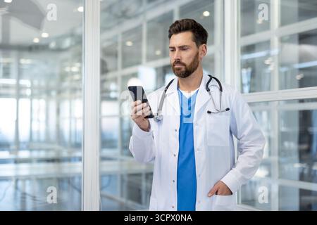 Medical professional standing in a modern clinic or hospital, intently looking at his mobile phone, connecting with patients through digital health technology Stock Photo