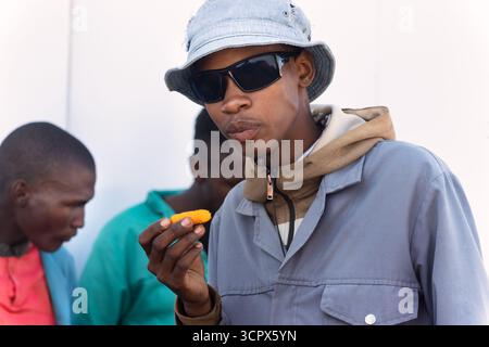 african construction workers, lunch break eating corn chips, sited outdoors , in the yard of a warehouse in workwear uniform with hat and sunglasses s Stock Photo
