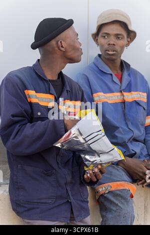 two african workers, friends talking and eating chips, sited outdoors in the yard of a warehouse in workwear uniform standing in front of a warehouse Stock Photo
