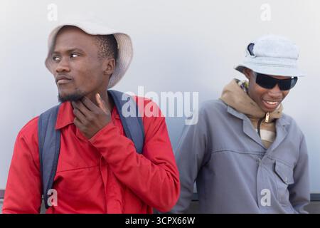 two african workers, sited outdoors in the yard of a warehouse next to building wall, wearing beanie and sunglasses Stock Photo