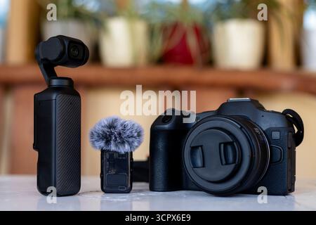 Handheld Gimbal Camera, Wireless Microphone and Interchangeable Lens Mirrorless Digital Camera on White Table with Blurred Potted Plants Background Stock Photo