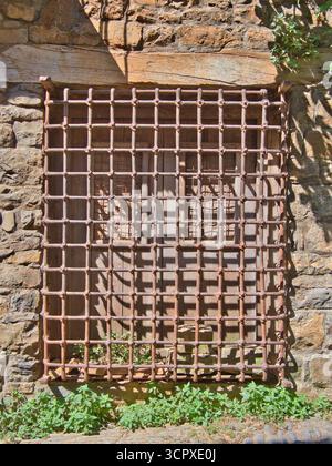 Rustic Window with Wrought Iron Grate - Close-up of an old wooden window with a rusty square wrought iron security grate set into a rough, historic st Stock Photo