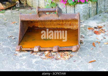 A rusty excavator bucket sits on a paved surface, filled with orange water and surrounded by fallen leaves. Stock Photo