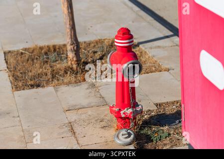 A bright red fire hydrant stands on a tiled surface, next to a tree and a red structure with white shapes. Stock Photo