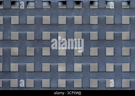 Close-up of a gray metal grate with square openings and a gray background surface featuring tiny red and white specks. Stock Photo