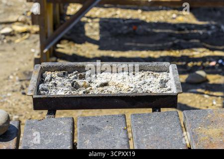 Close-up of a rectangular metal ash tray filled with ash and charcoal resting on top of a metal structure, set outdoors. Stock Photo