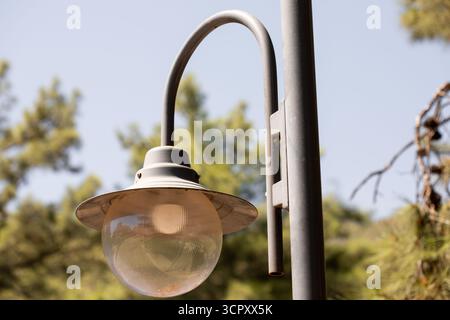 A close-up shot of a gray metal street light with a round glass globe, set against a backdrop of trees and a blue sky. Stock Photo