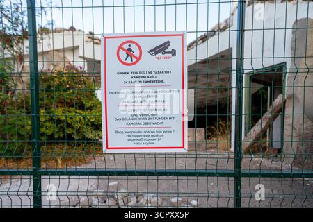 A warning sign prohibiting entry is posted on a green wire fence in front of a derelict building. Stock Photo